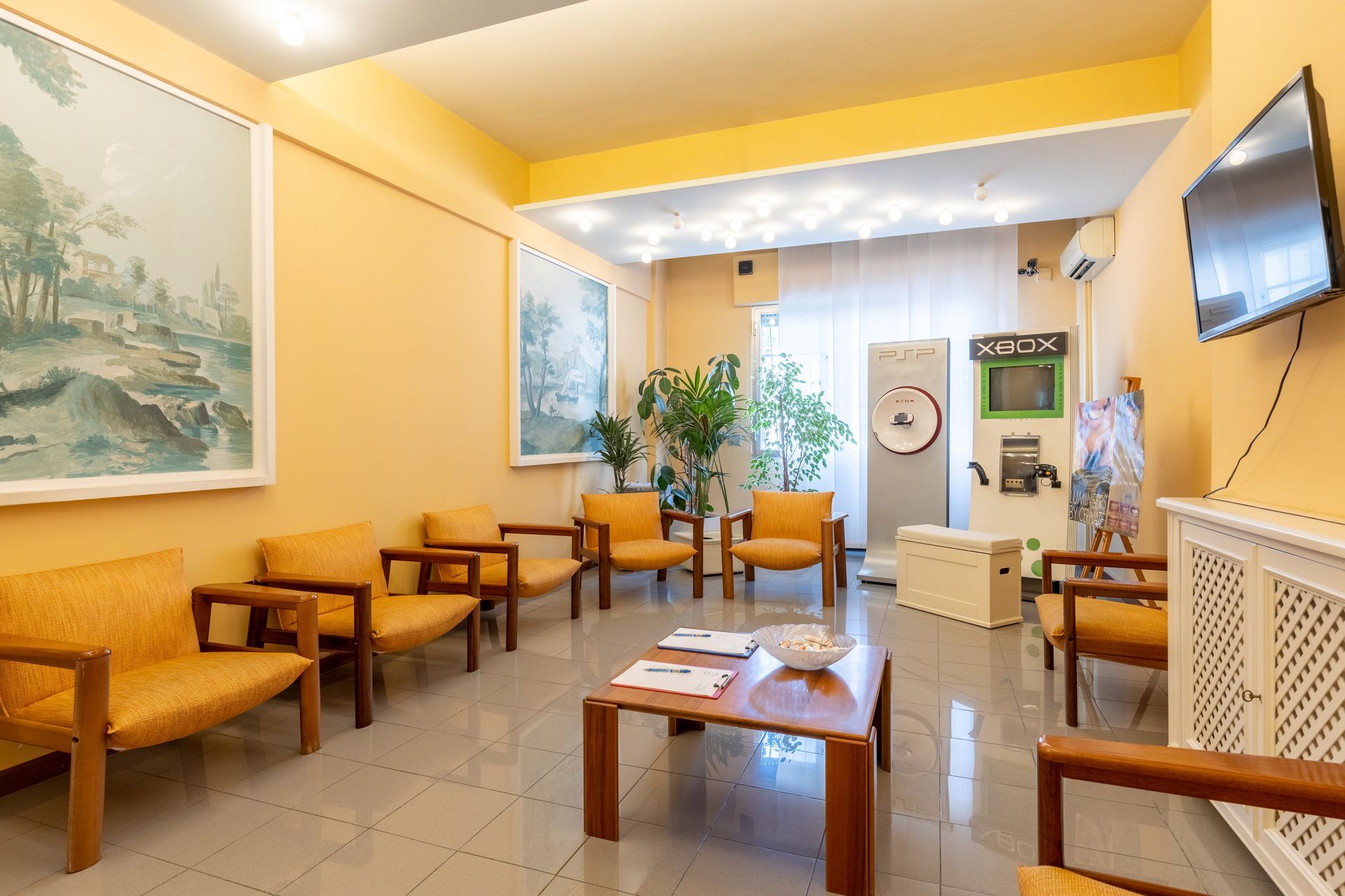Luminosa sala d'attesa con sedie in vimini, tavolino da caffè, piante, quadri e televisore a parete.