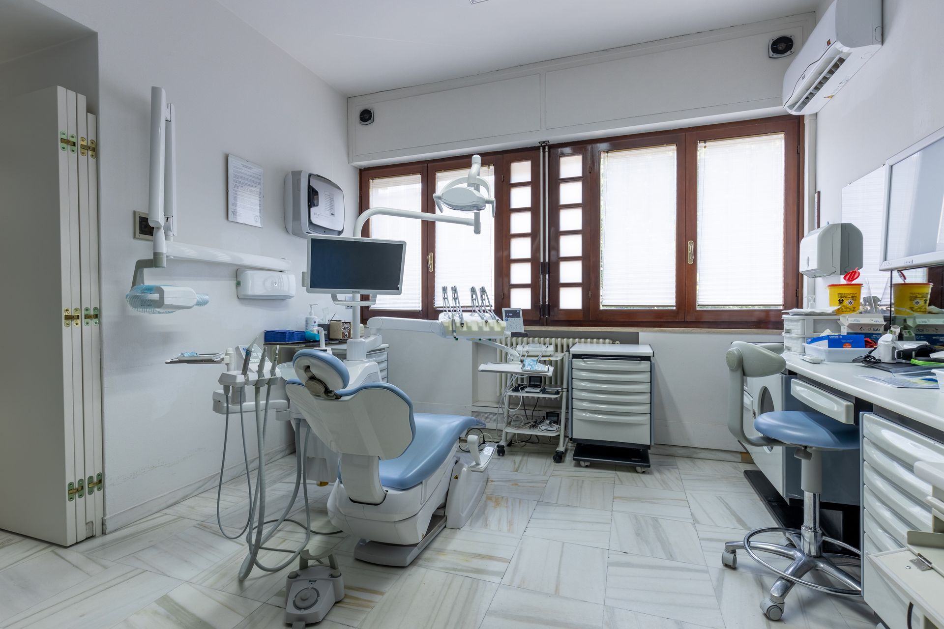 Studio dentistico vuoto con poltrona, strumenti, armadietti e grandi finestre in una stanza bianca e luminosa.