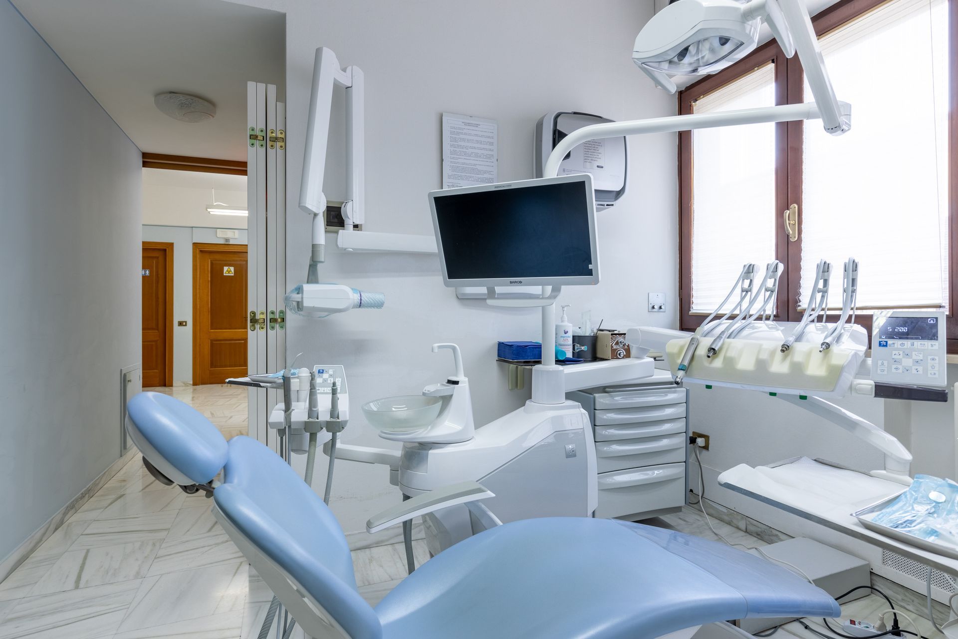 Sala per visite odontoiatriche con poltrona blu, monitor e strumenti dentali vicino a una finestra.
