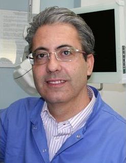Un uomo con camice blu e occhiali è in piedi in un ambiente clinico, con un monitor alle sue spalle.