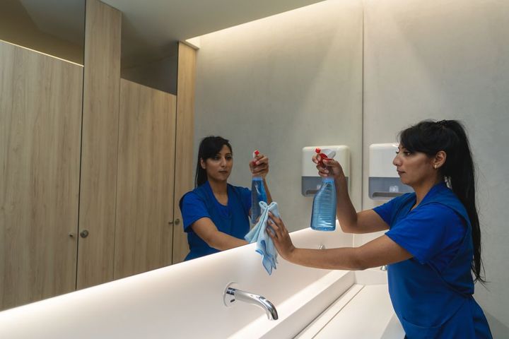 A woman is cleaning a bathroom mirror with a cloth and spray bottle.
