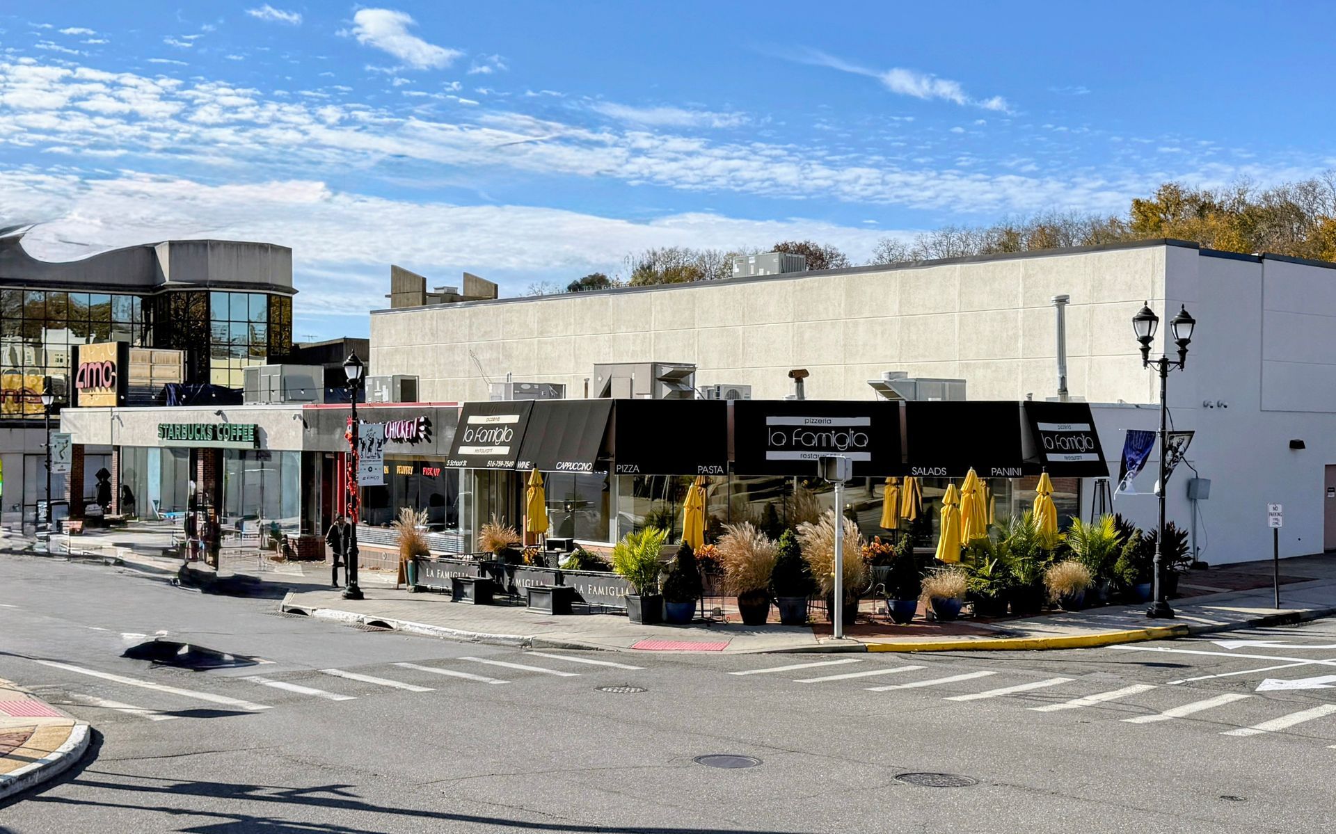A sunny street corner features a restaurant with an outdoor patio, yellow umbrellas, and black awnings against a tan wall.