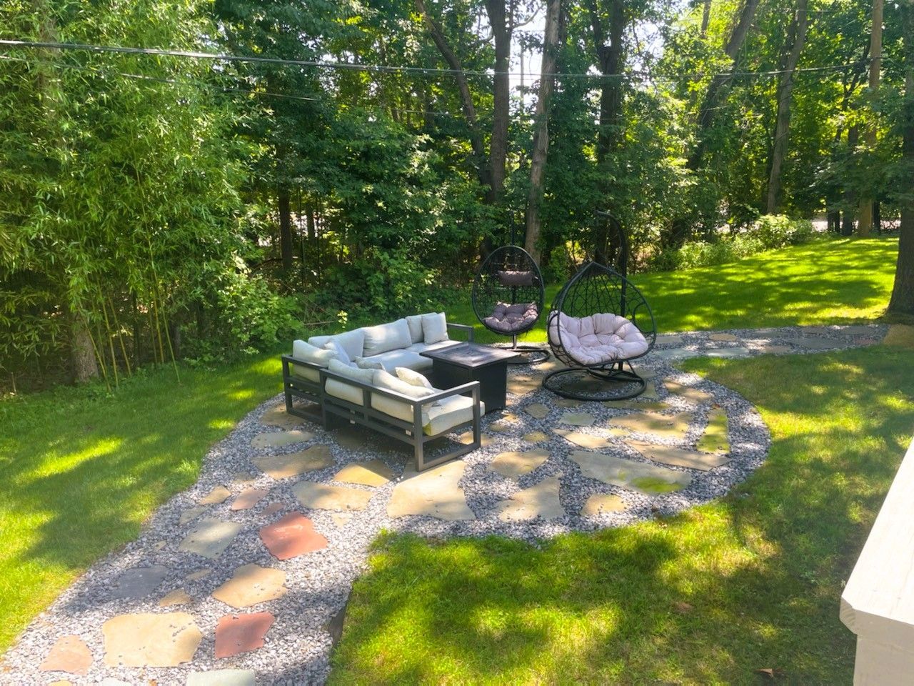 Outdoor patio with a grey sofa and two hanging chairs on a stone-and-gravel path in a grassy, wooded backyard.