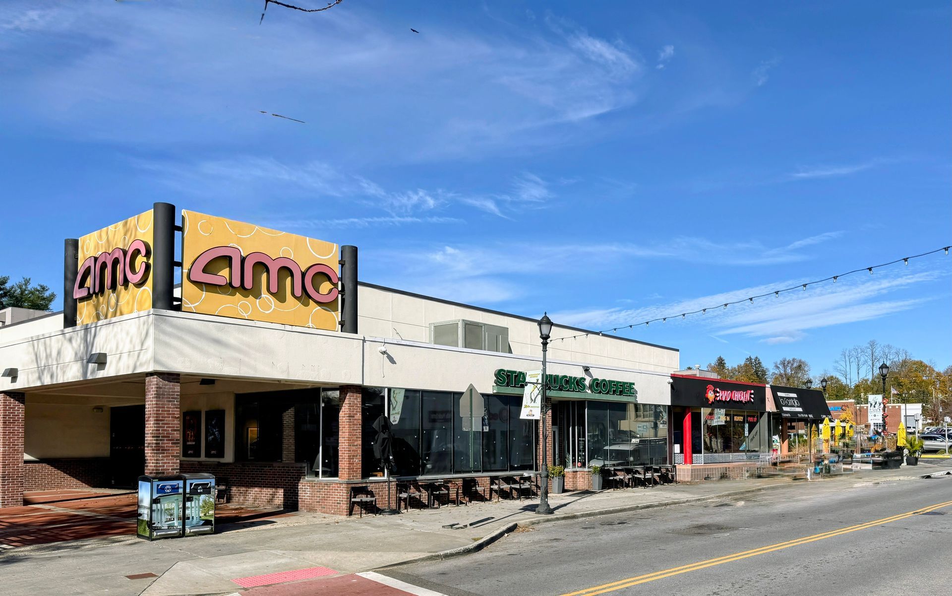 An outdoor street-level view of an AMC theater building and adjacent storefronts under a clear blue sky.