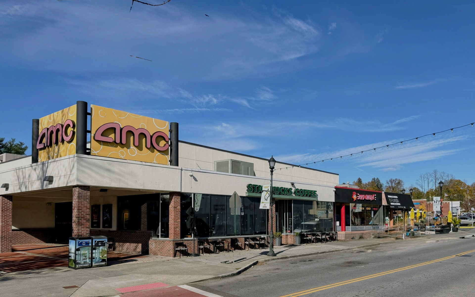 An AMC movie theater and adjacent storefronts along a road under a bright blue sky.