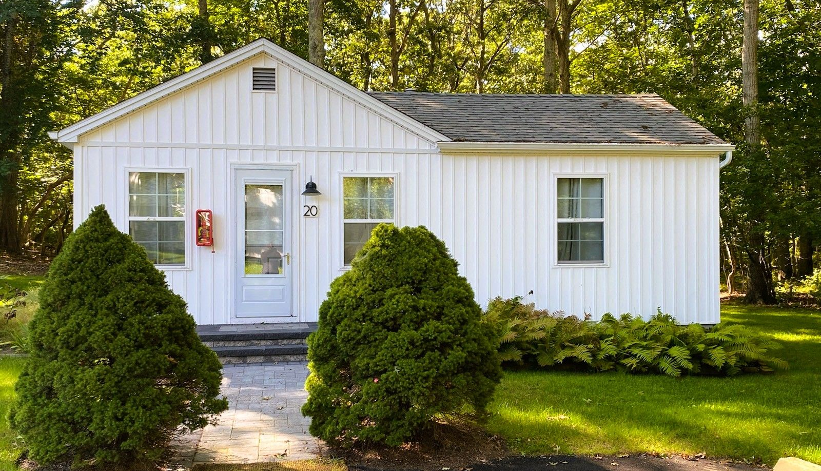 A small, white, single-story cottage with vertical siding, surrounded by trees and two large, cone-shaped green shrubs.