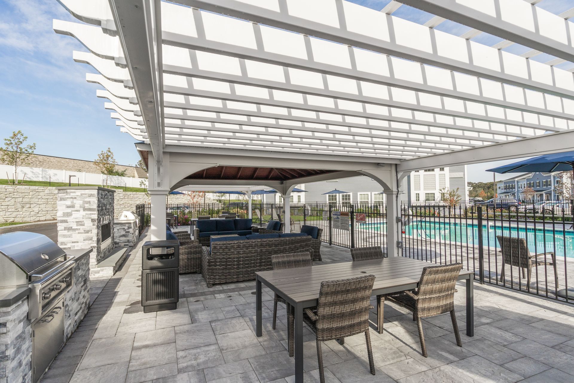 A patio with a pergola, dining table, lounge seating, and a grill, adjacent to an outdoor swimming pool.