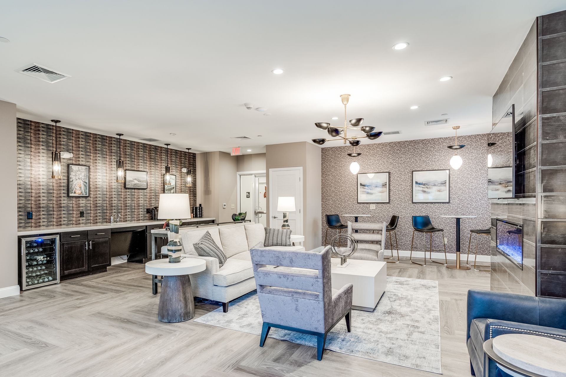 Modern apartment community lounge with a seating area, kitchenette, beverage fridge, and decor featuring textured walls.