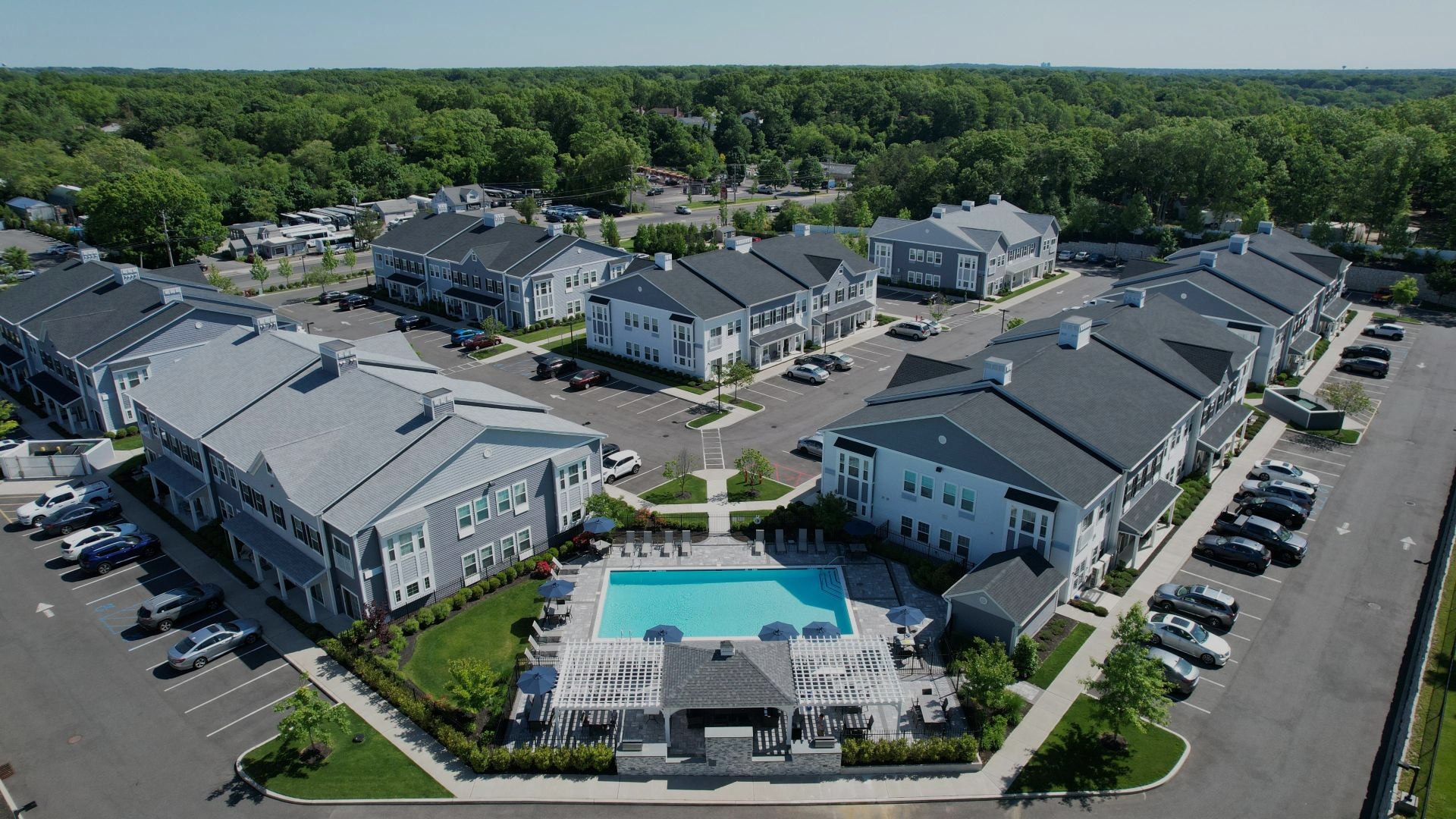 Aerial view of a residential apartment complex featuring multiple grey buildings, a swimming pool, and parking lots.