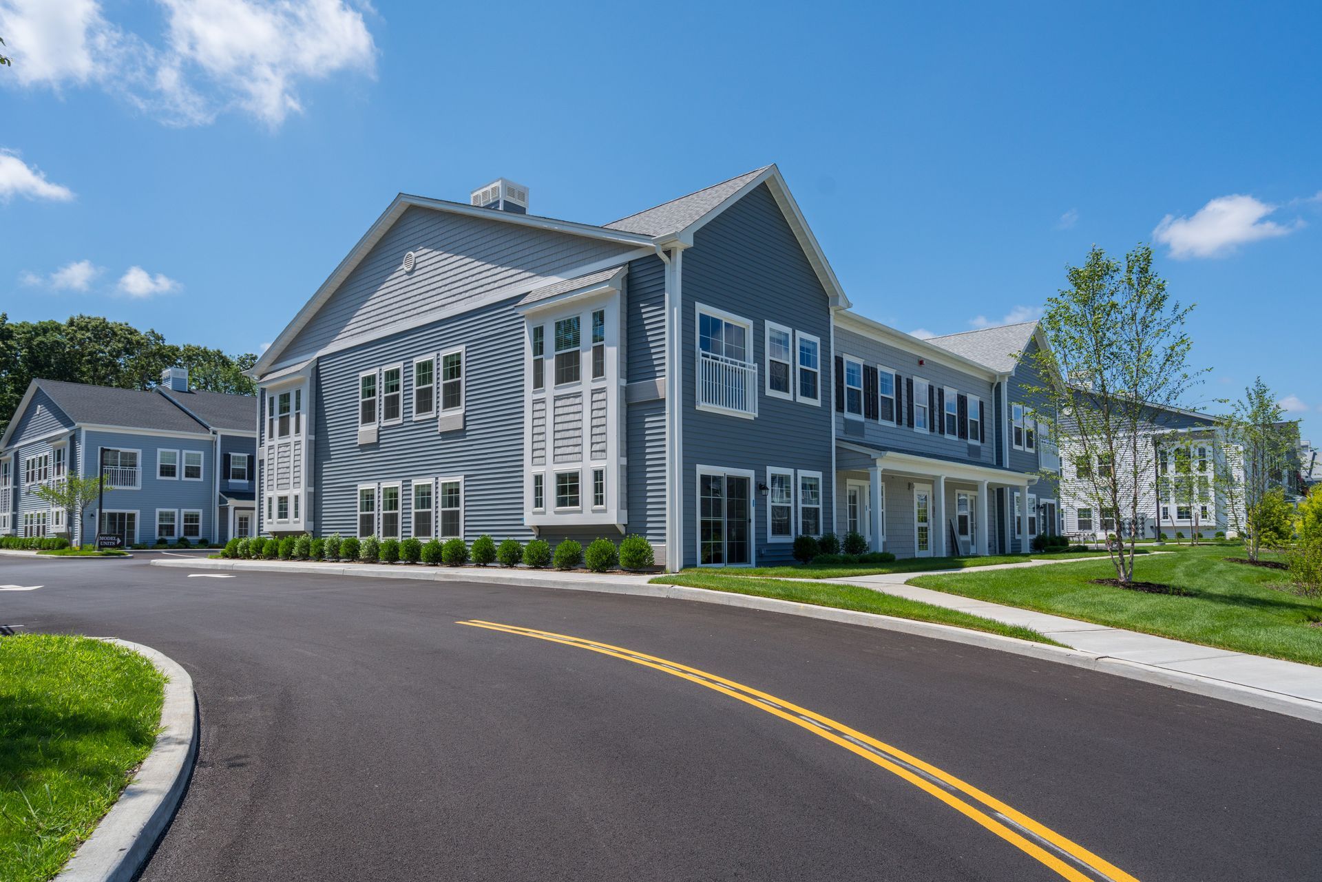 A modern, two-story apartment complex with blue siding, a grey roof, and a curved asphalt road in the foreground.
