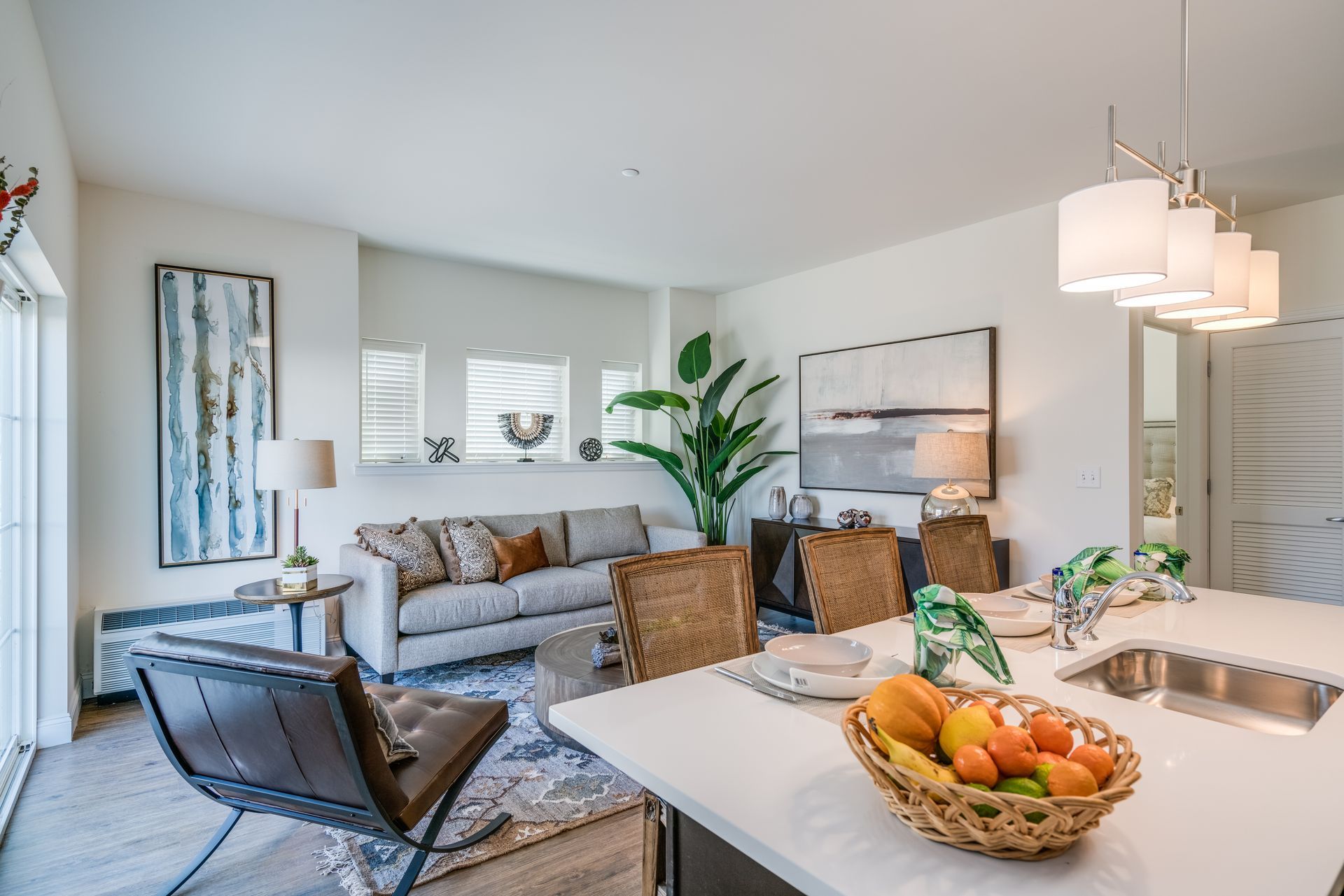 A modern, open-concept apartment interior showing a kitchen island with fruit, a living area with a sofa, and decor.