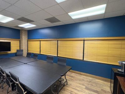 Empty conference room with blue walls, slatted blinds, folding tables, and stacked chairs under ceiling lights