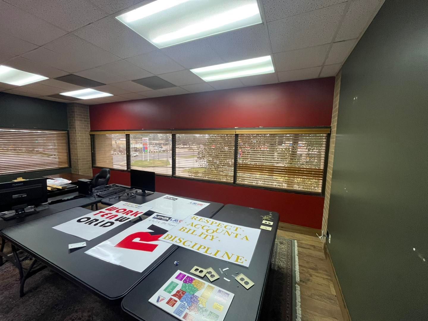 Office meeting room with signs and posters spread across tables, red walls, and window blinds.