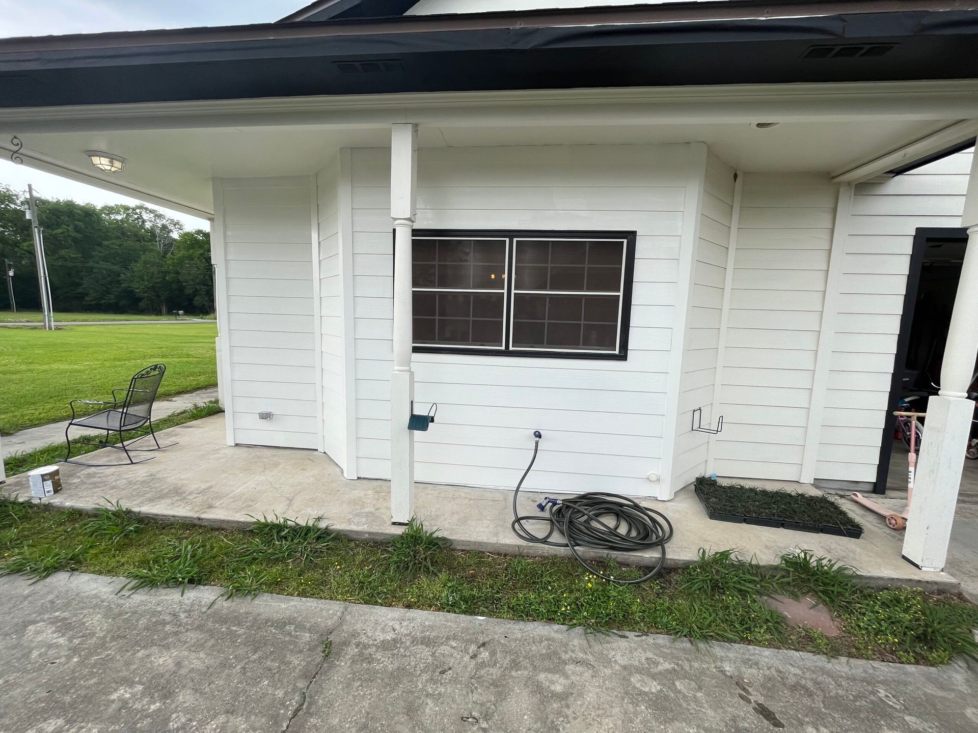 White house exterior with a small window, porch posts, concrete driveway, and coiled hose by the wall