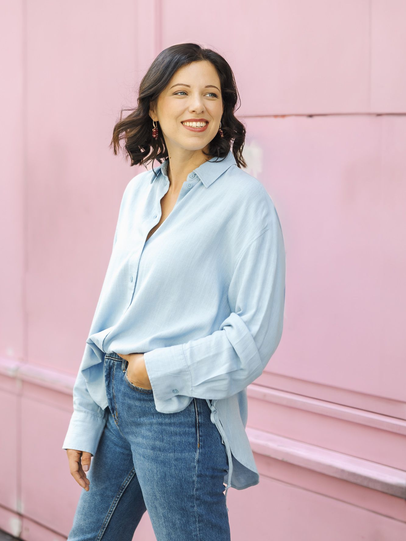 Eine Frau in hellblauem Hemd und Jeans lehnt lächelnd im Freien an einer rosa Wand.