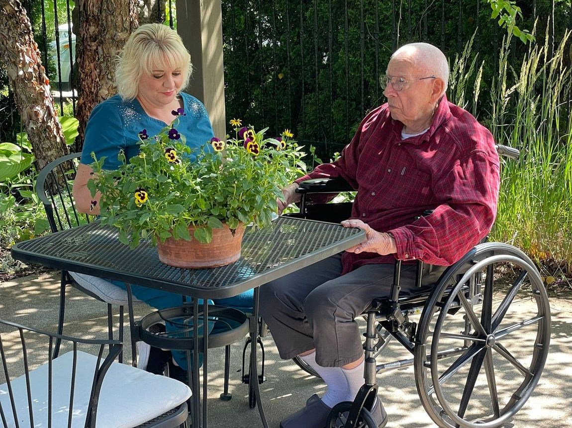 Woman and man in a wheelchair sit at a table with potted flowers outside. Man grips table, woman looks at flowers.