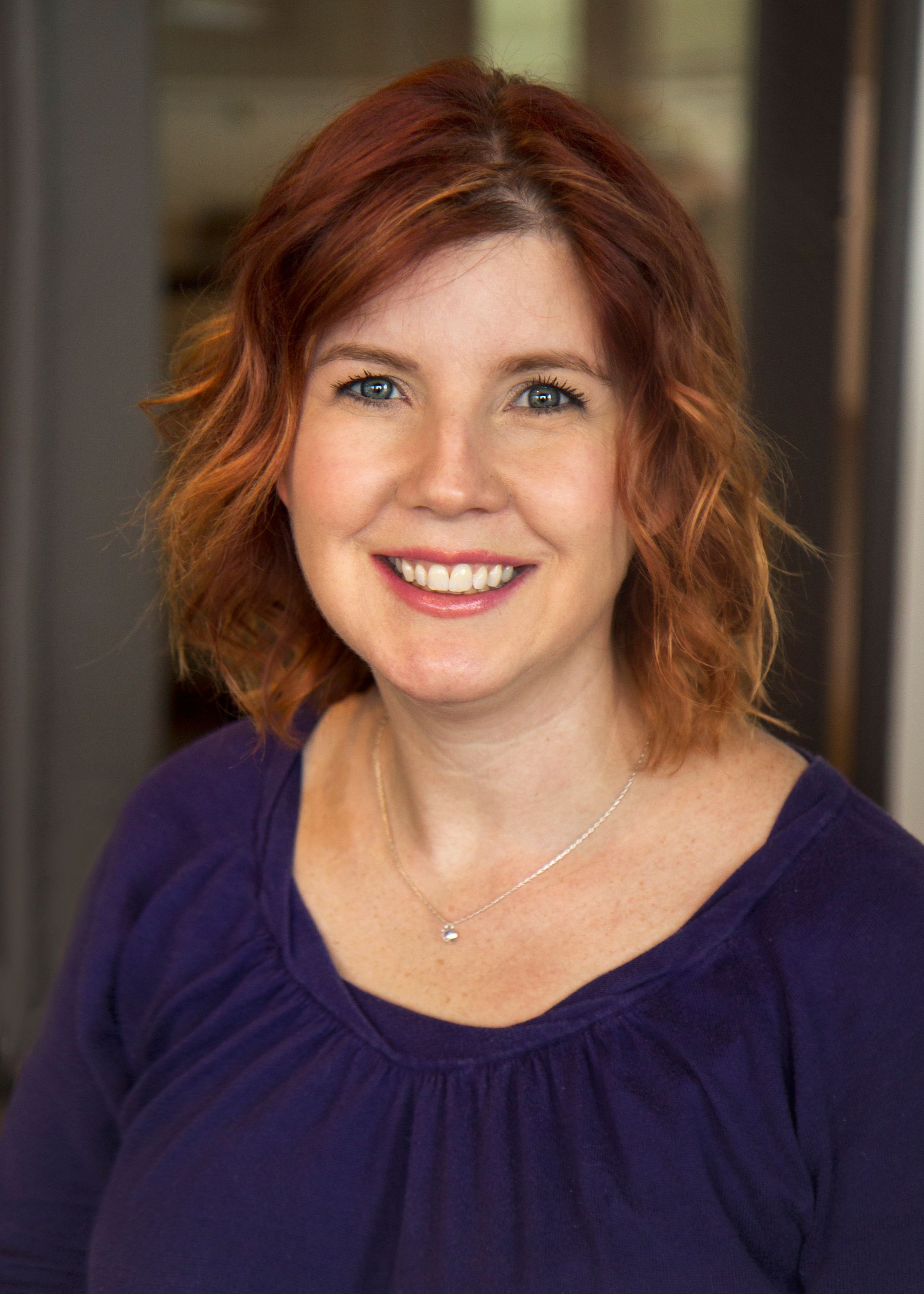 Woman with red hair, smiling. Wearing a purple shirt and small necklace. Blurred background.