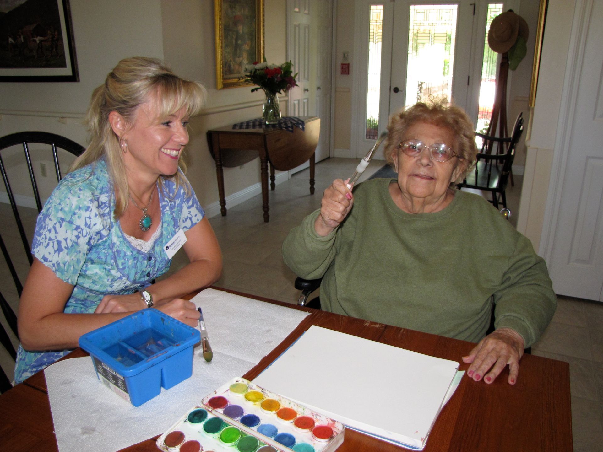 Woman assists elder with watercolor painting. Supplies on table, they smile, sunny room.