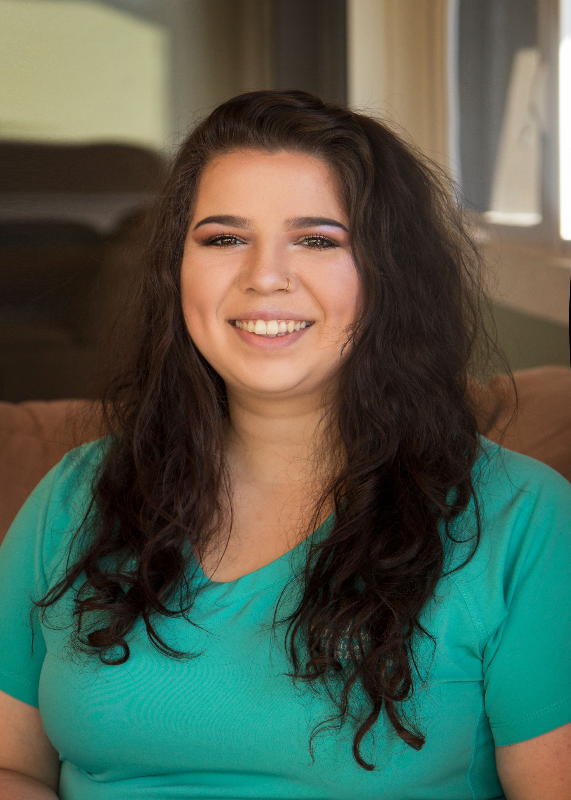 Woman with long, wavy dark hair smiles at the camera, wearing a teal v-neck top, sitting indoors.