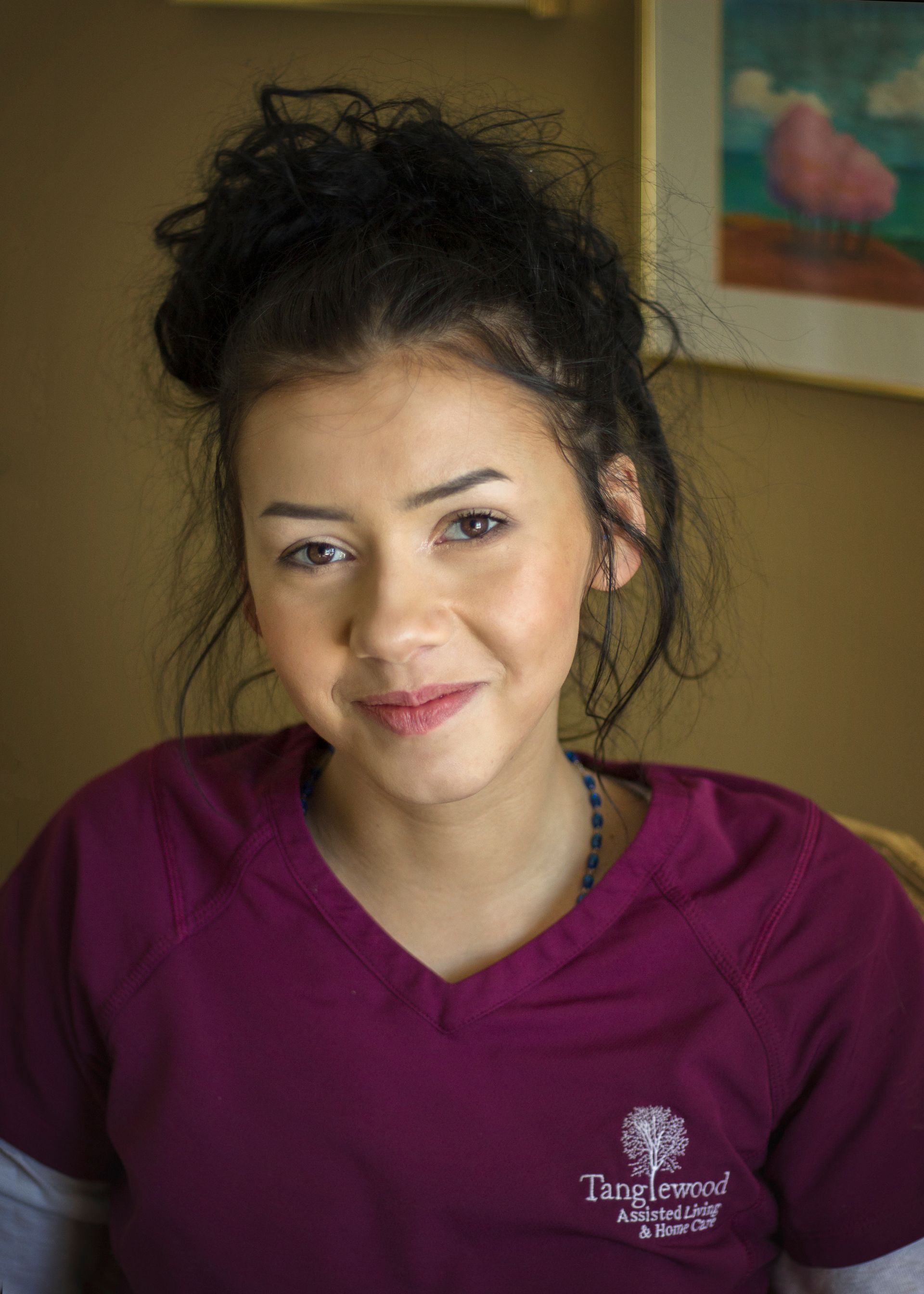 Woman with dark curly hair in burgundy scrubs, smiling, with a logo on the shirt.