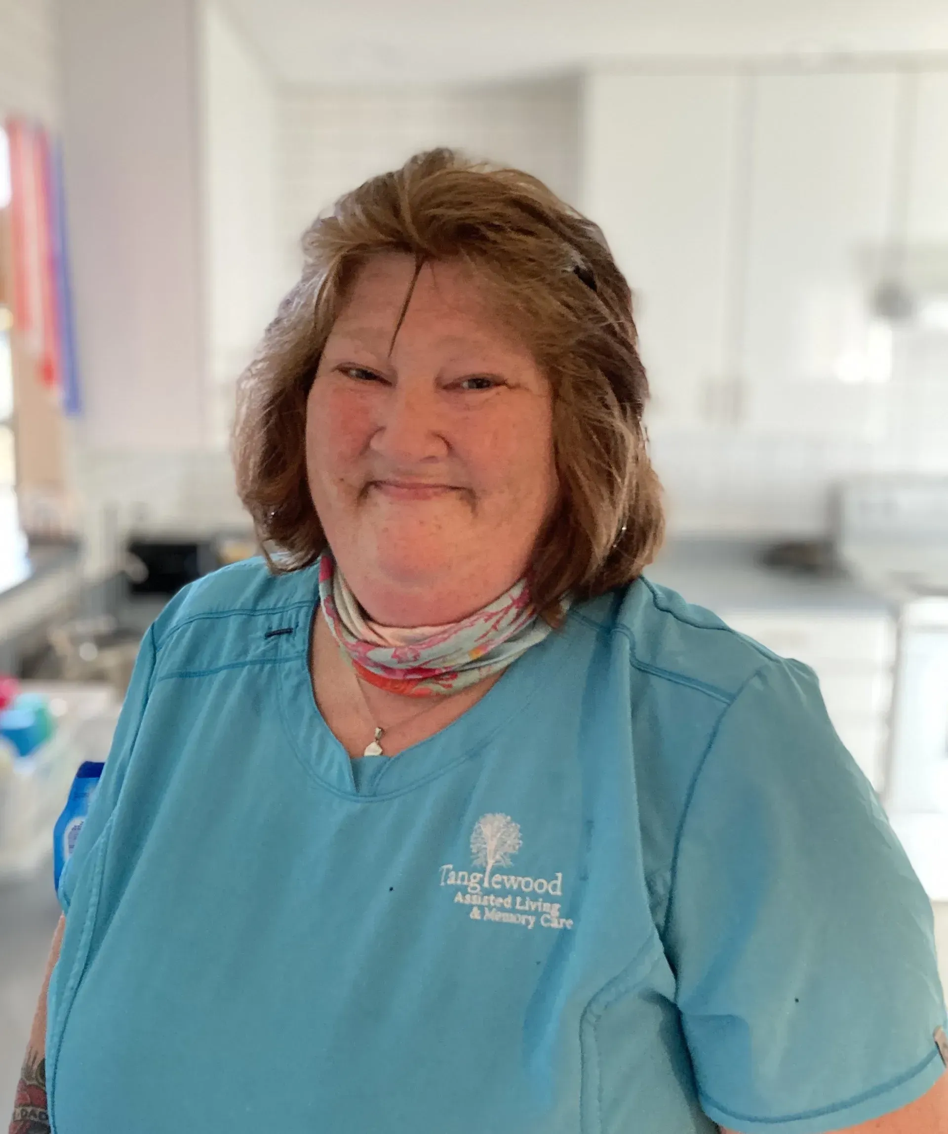 Woman in blue scrubs, smiling, indoors. A light blue shirt with a floral patterned neck scarf.