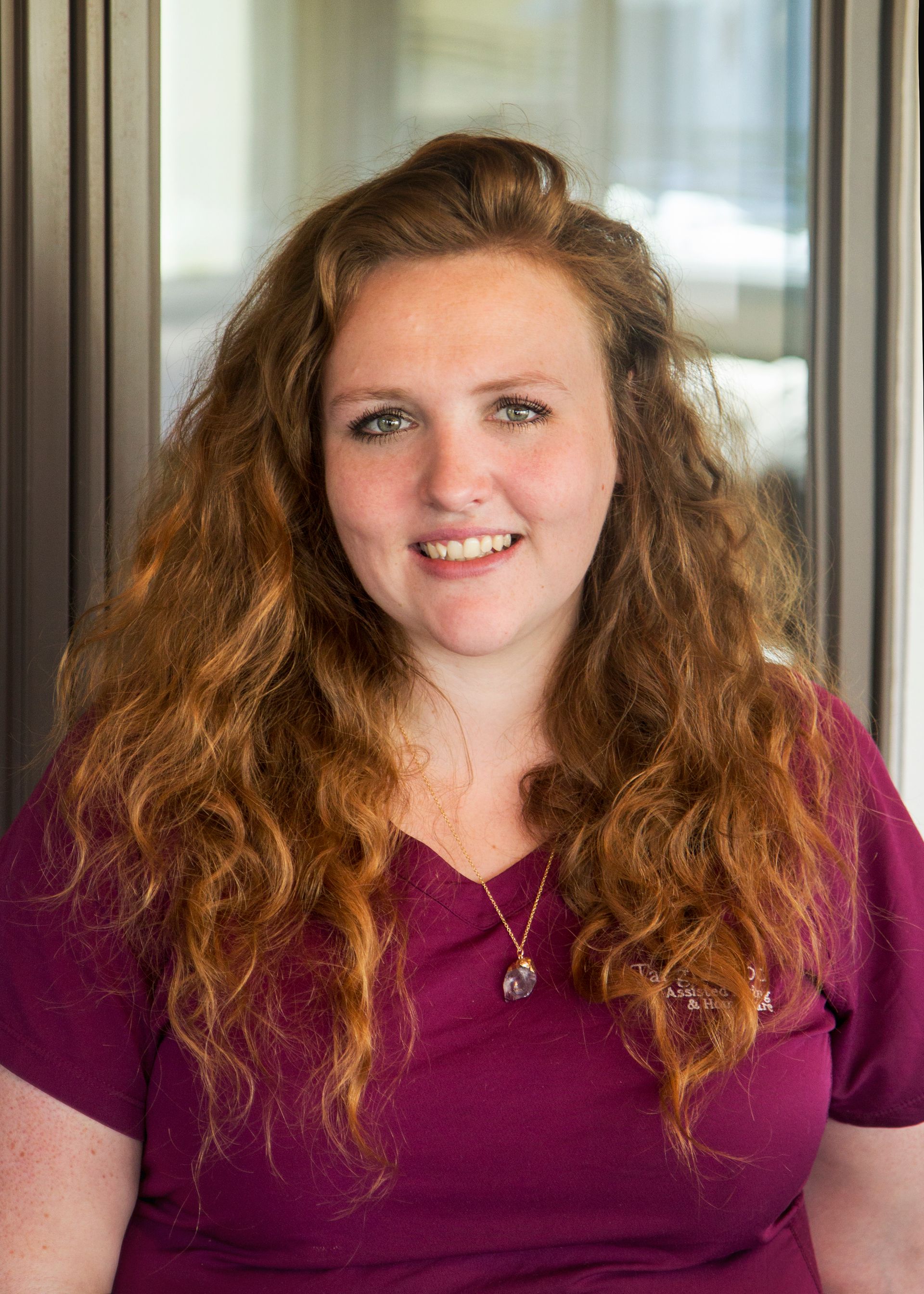 Woman with auburn curly hair smiles, wearing a purple shirt, near a window.