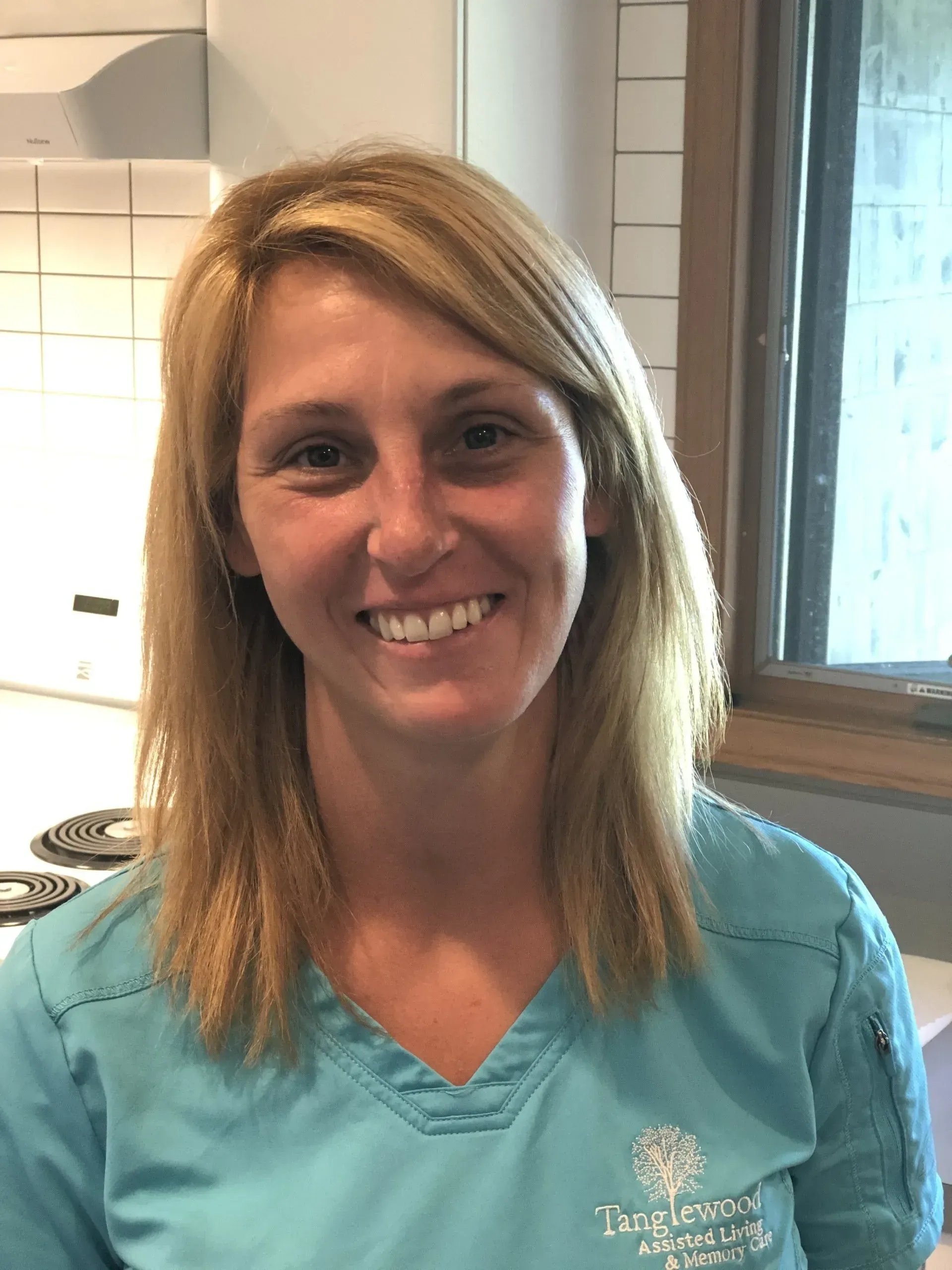 Woman with blonde hair wearing a teal scrub top smiles. Kitchen background.