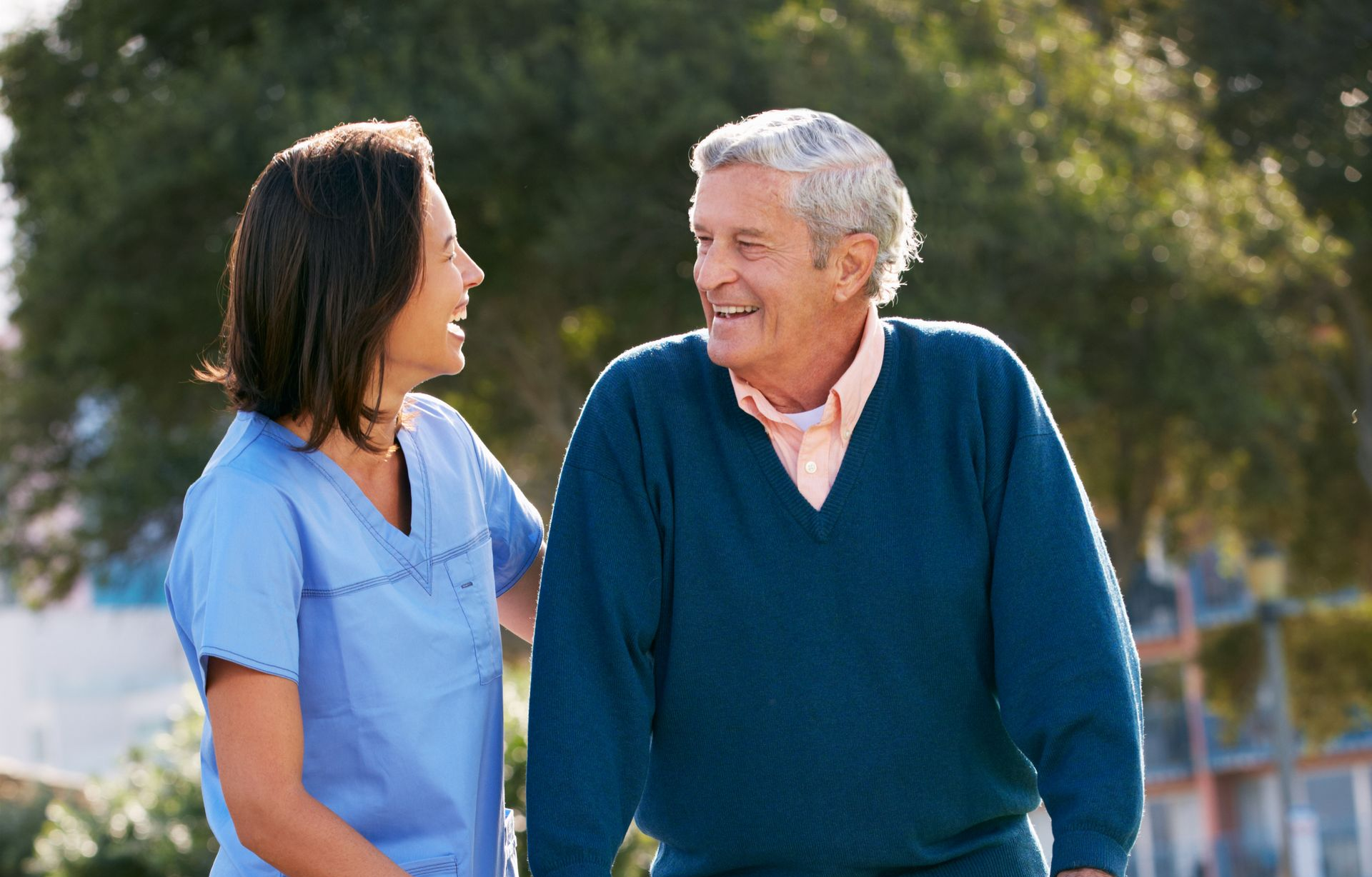 Woman in blue scrubs assists older man walking outdoors, smiling.