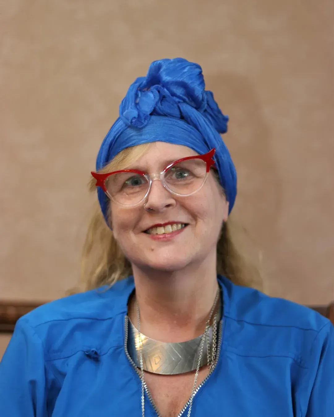 Woman wearing blue headscarf, glasses, silver necklace, and blue top. Smiling in front of a tan wall.