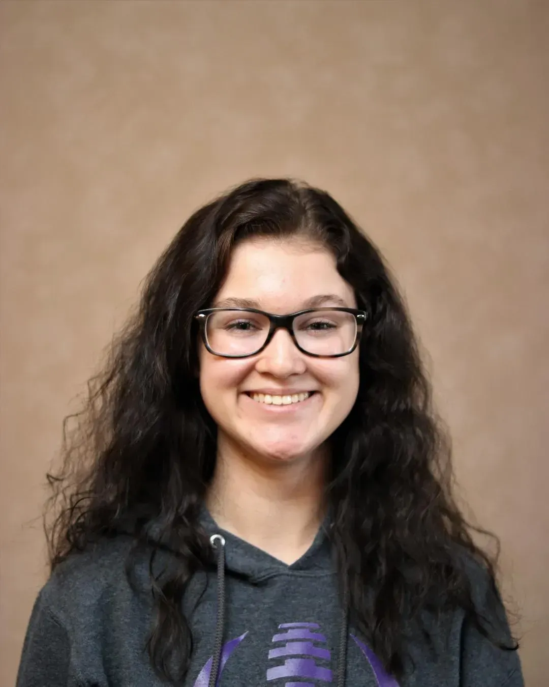 Woman with long dark curly hair, wearing glasses and a gray hooded sweatshirt, smiling. Beige background.
