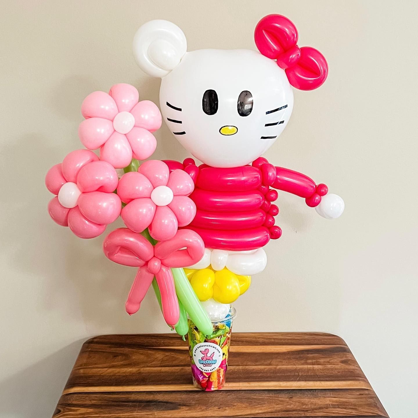 a hello kitty made out of balloons is holding a bouquet of pink flowers