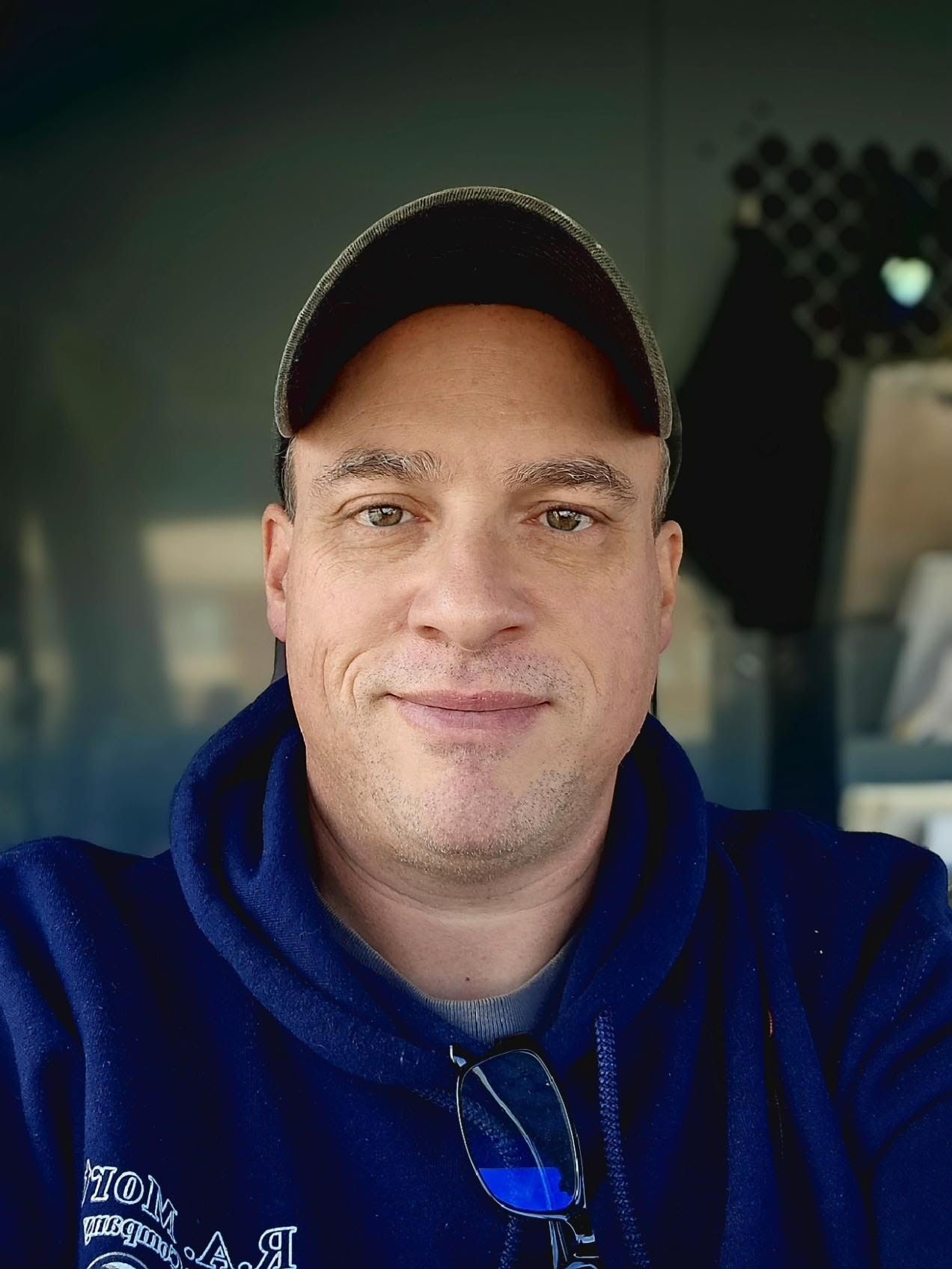A man wearing glasses and a blue sweater is smiling for the camera.