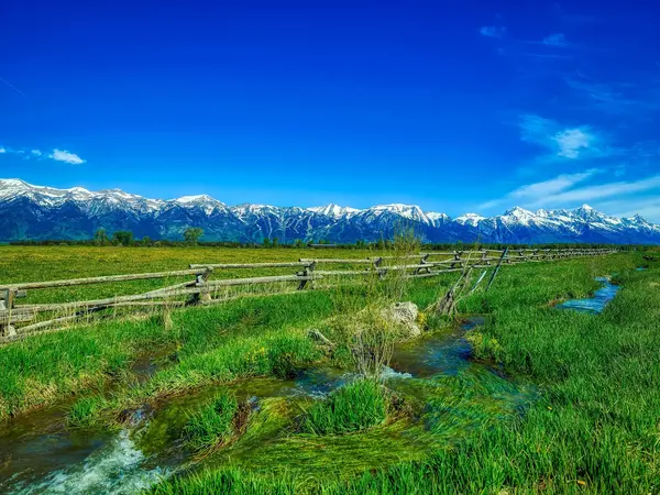 A wooden fence surrounds a grassy field with mountains in the background.