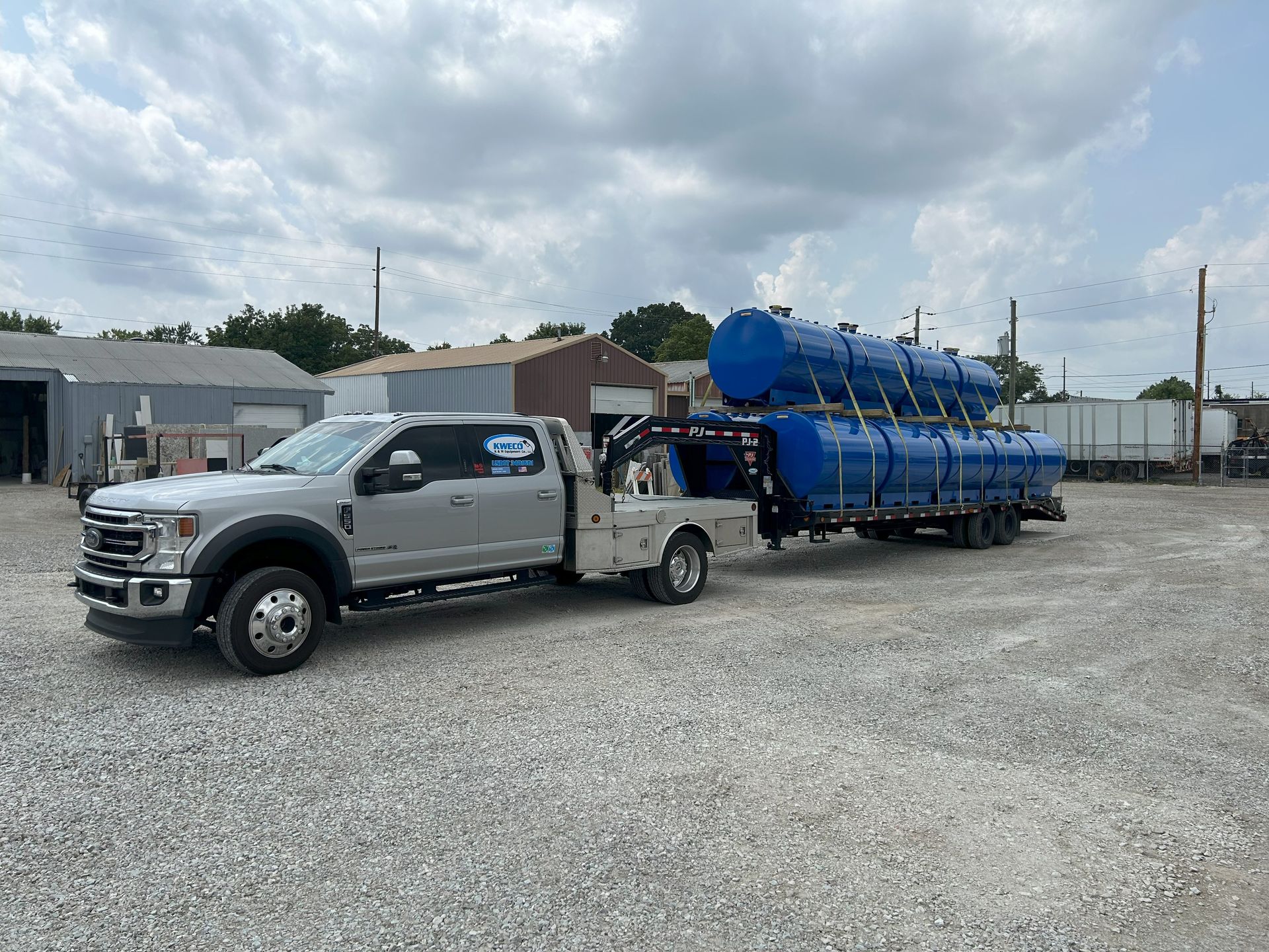 Silver pickup truck towing trailer with three blue tanks. Outdoors on gravel lot.