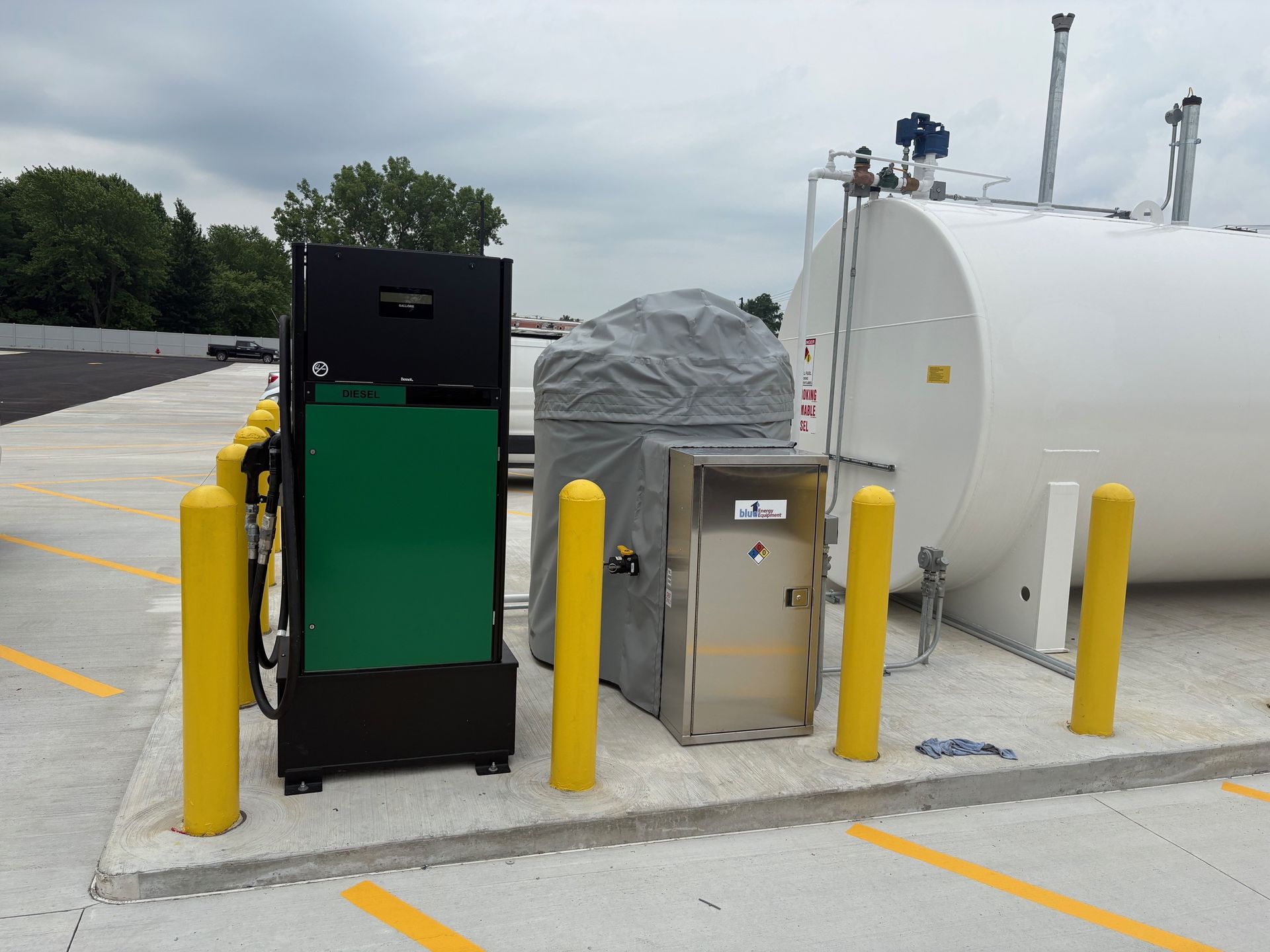 Fueling station with large white tank, green and black pump, and yellow bollards.