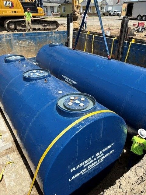 Two blue cylindrical tanks being installed in a construction site trench. Workers and machinery are visible.