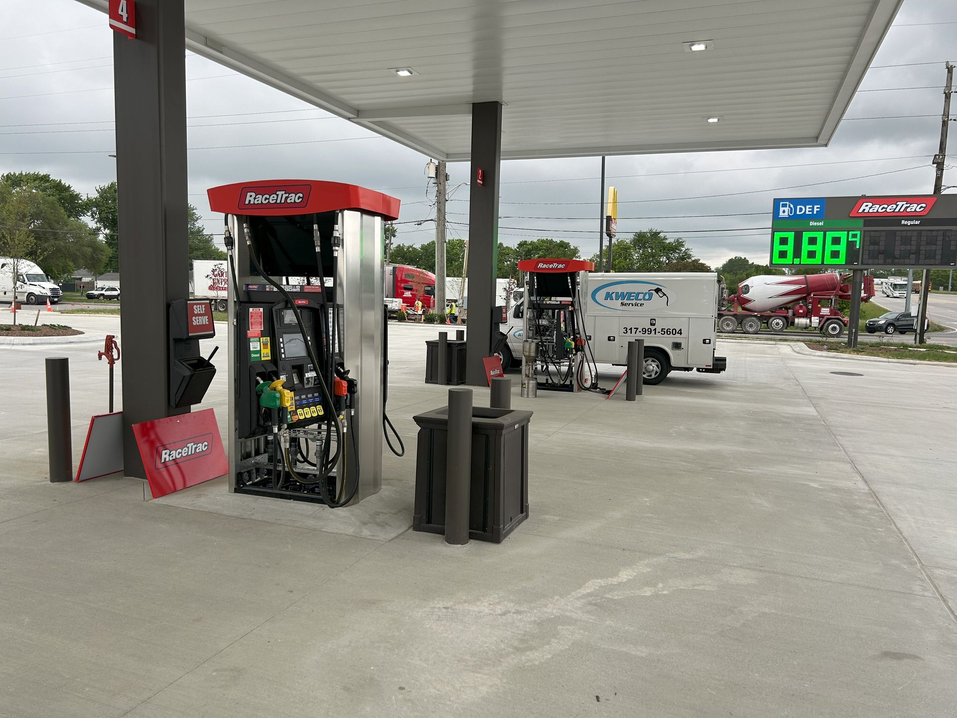 Gas station with red and gray fuel pumps under a canopy, green price display; white service truck.
