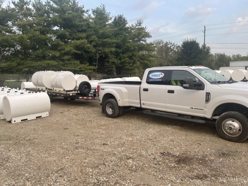 White truck towing a trailer loaded with white cylindrical tanks; outdoors, cloudy sky.