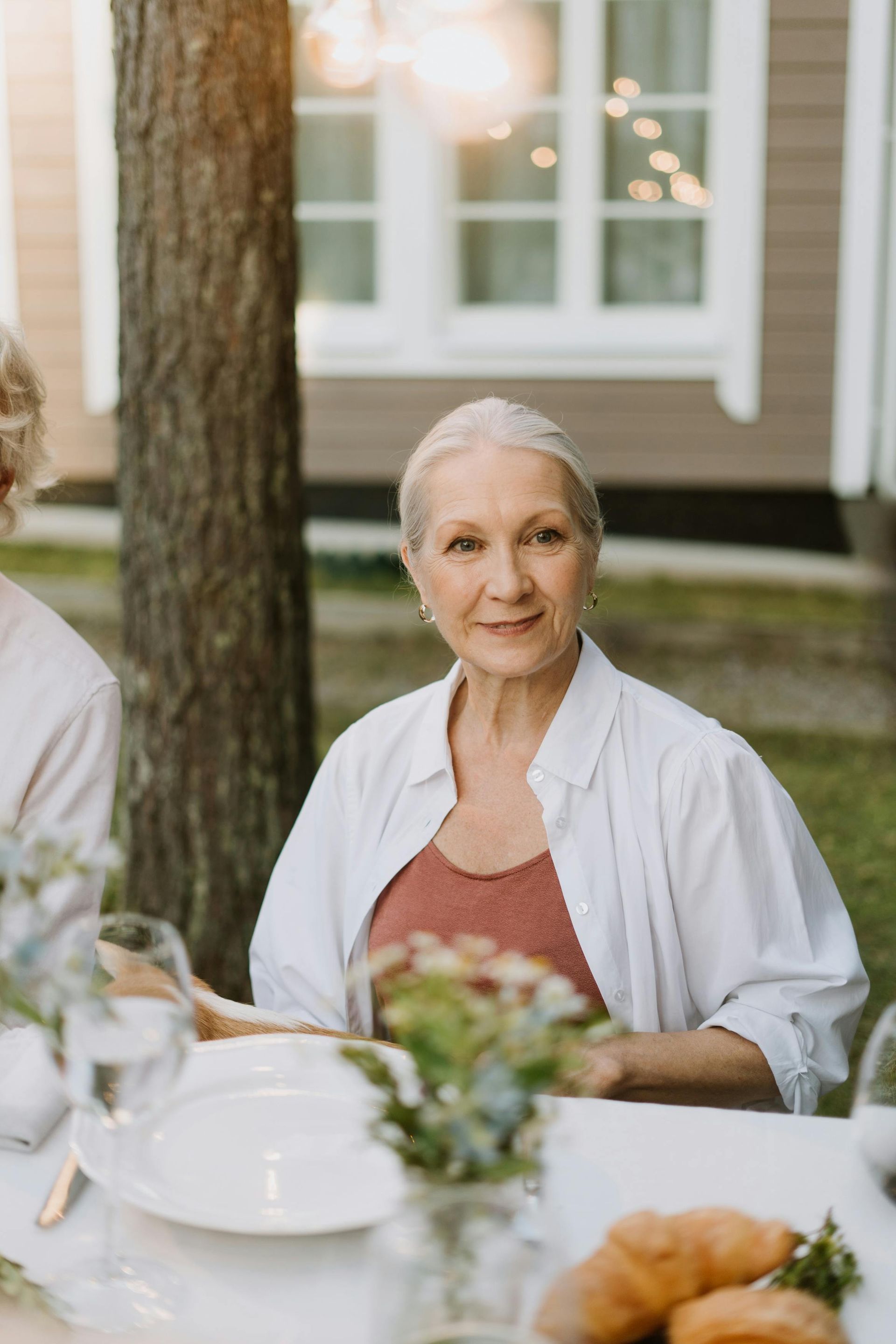 An elderly woman is sitting at a table with plates and glasses of wine.
