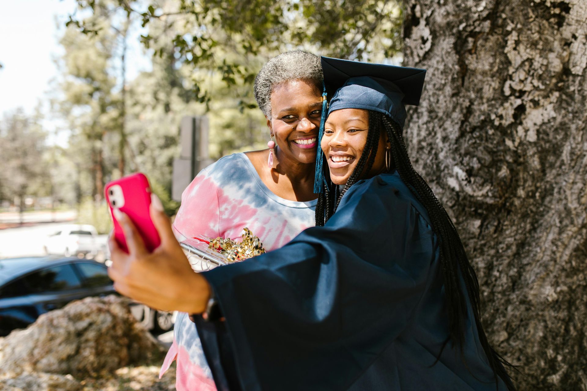 A woman in a graduation cap and gown is taking a selfie with her mother.