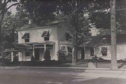 Black-and-white photo of a two-story house with awnings, trees, and a picket fence in front