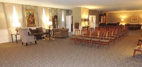 Bright lounge with armchairs, rows of chairs, and a piano in a carpeted room with curtains and paintings.