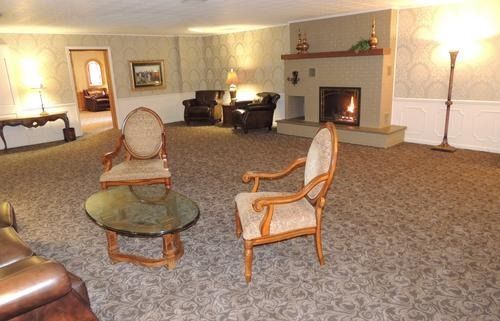Spacious beige living room with patterned carpet, fireplace, piano, and two chairs by a glass coffee table