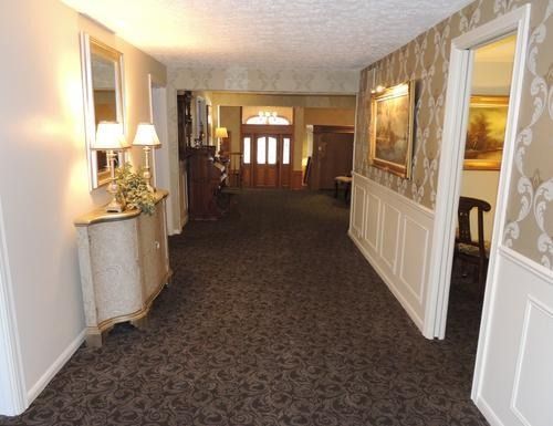 Carpeted hotel hallway with framed art, wall sconces, and white wainscoting leading to double doors.