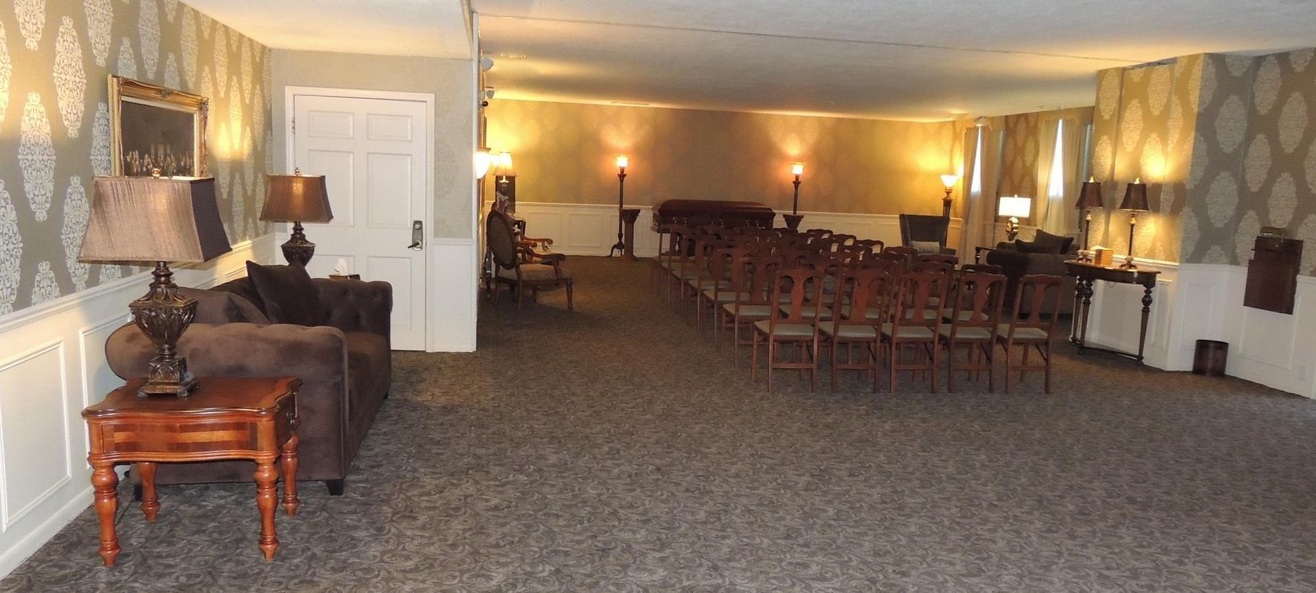 Wide banquet hall with rows of chairs, tables, and warm wall lighting on carpeted floor