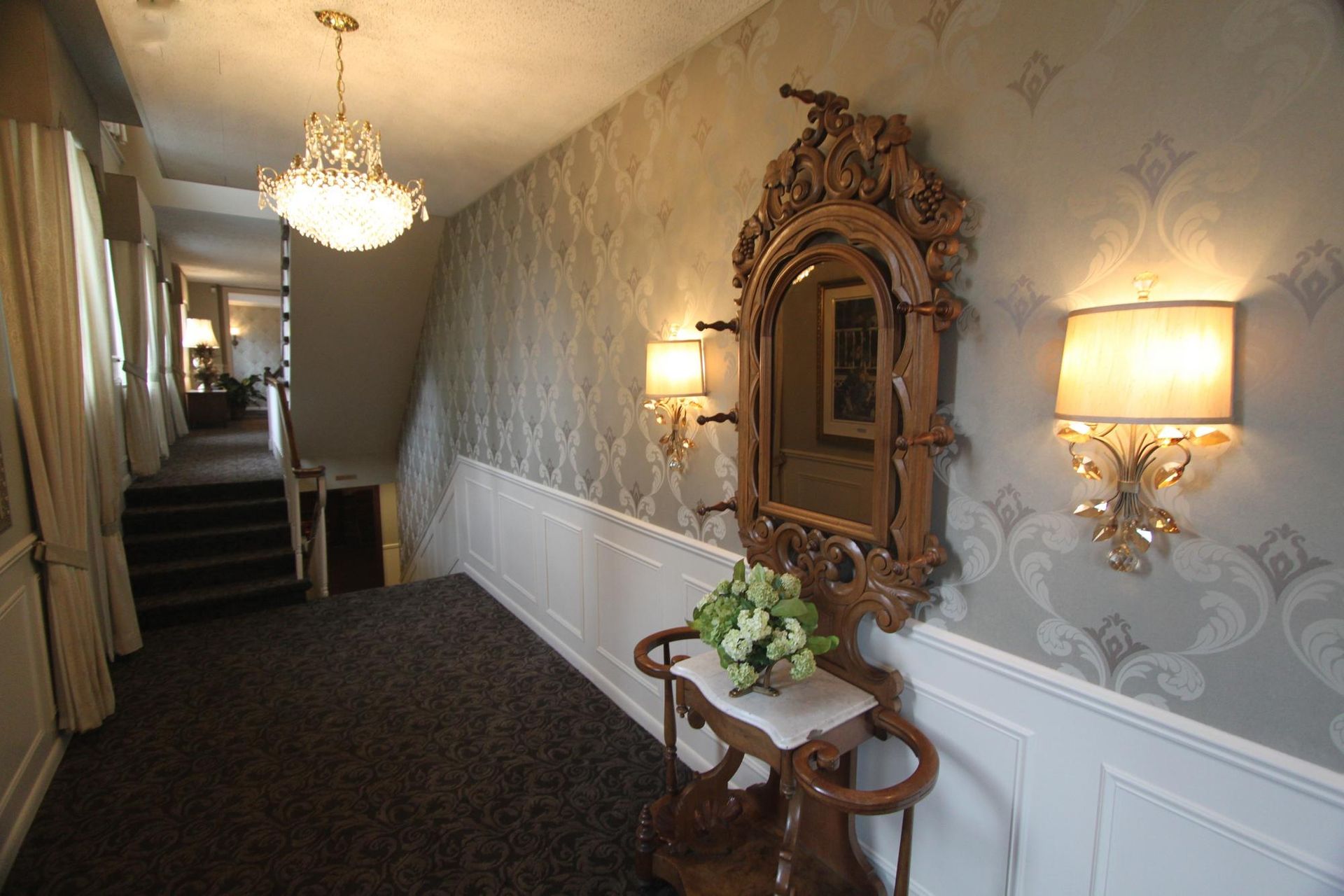 Elegant hotel hallway with ornate mirror, wall sconces, chandelier, and dark carpet leading to stairs