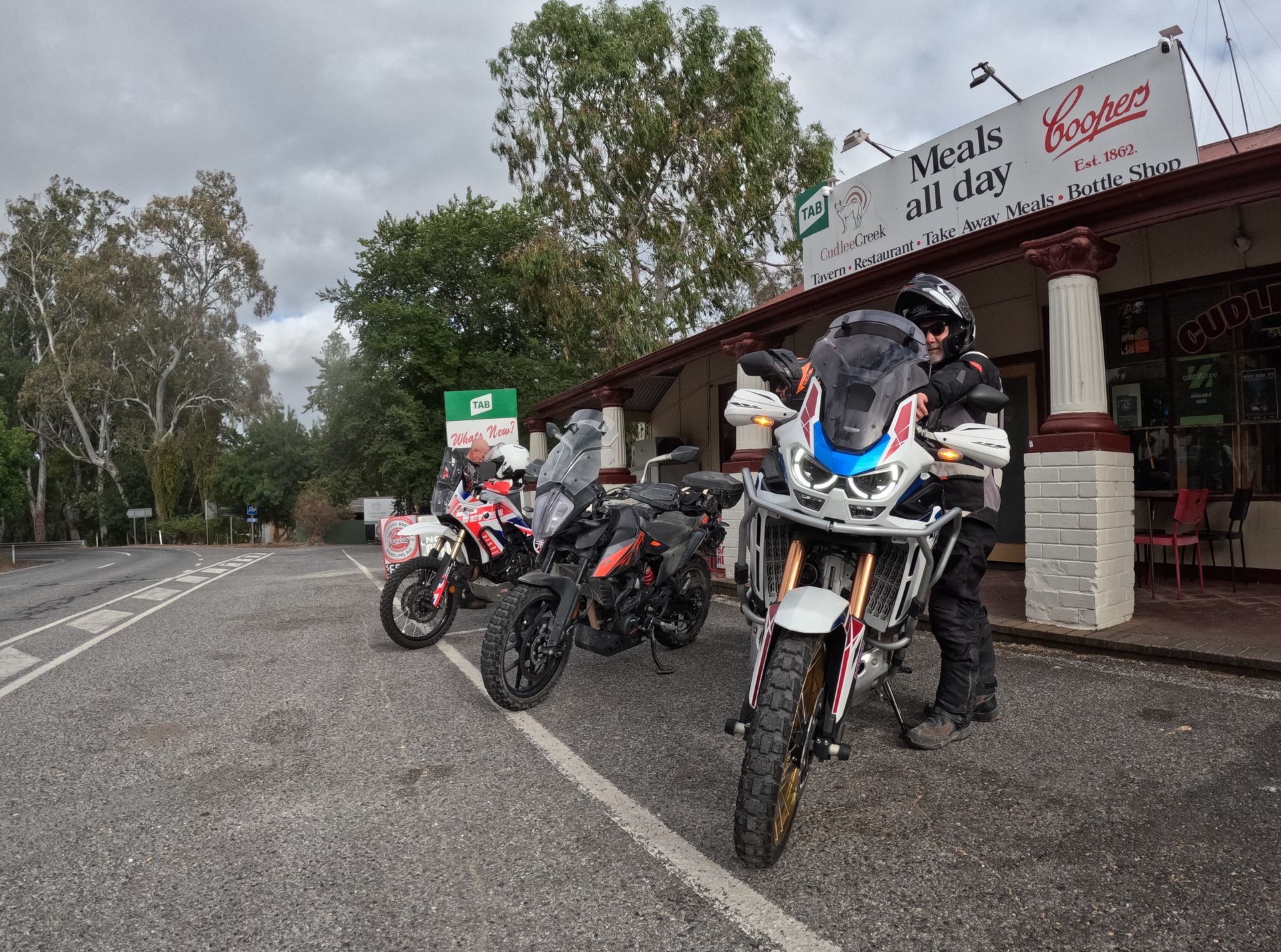 Motorcycles parked by a roadside café, with riders in helmets beside them on an overcast day