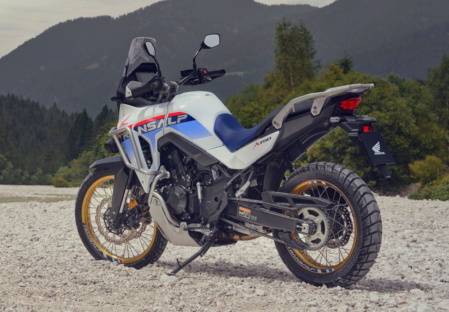 White and blue adventure motorcycle parked on gravel with forested mountains in the background