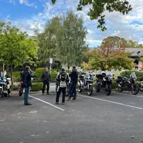 People standing near parked motorcycles in a tree-lined parking lot under a partly cloudy sky.
