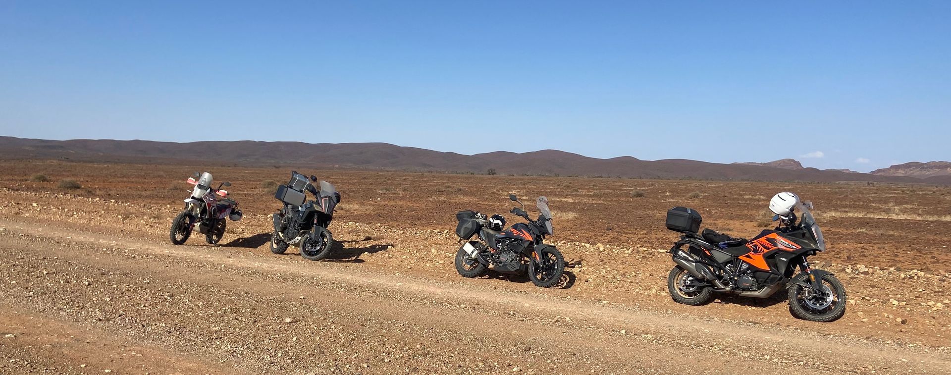 Four motorcycles parked on a dusty desert road with riders beside them under a clear sky